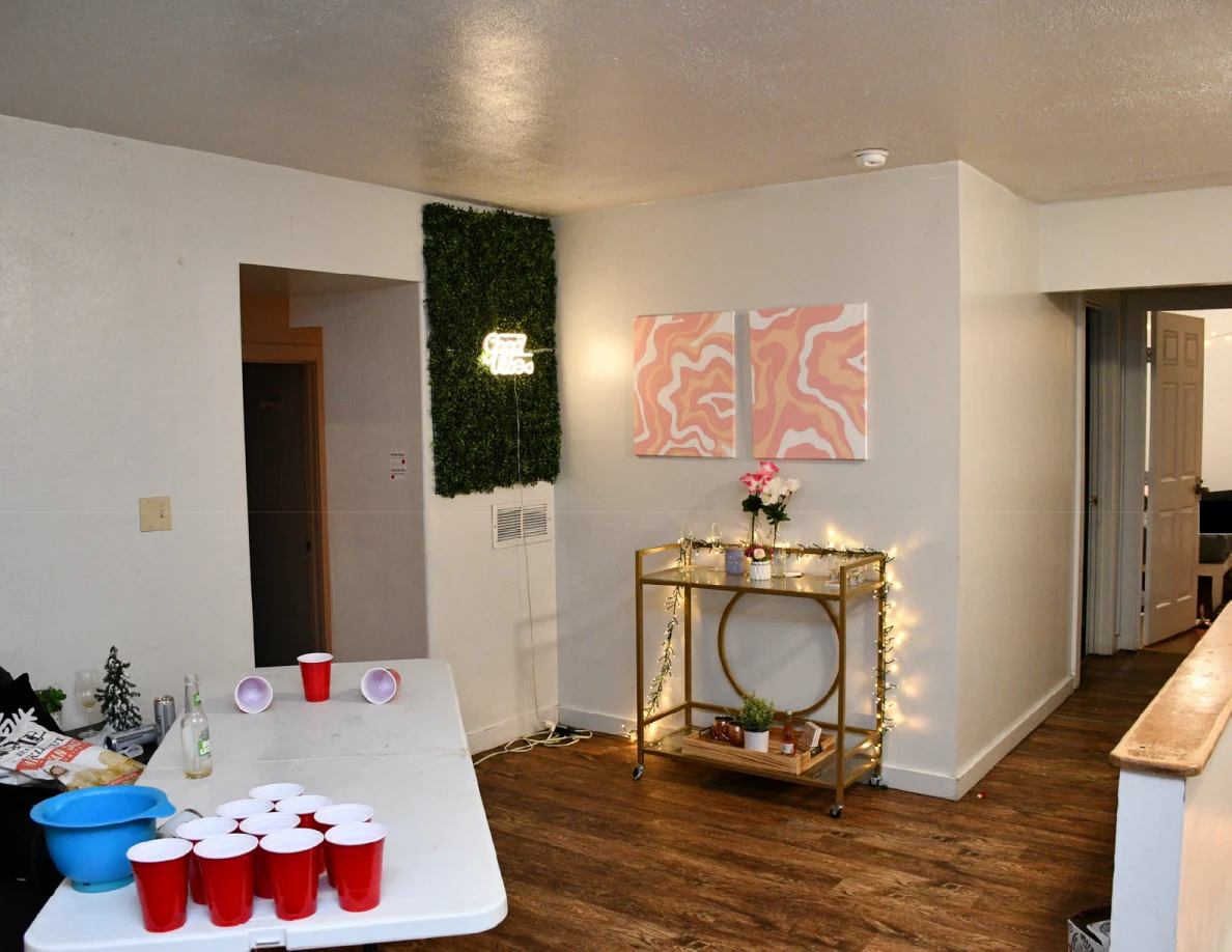 White cup-pong table with red cups beside a gold drinks cart, pink wall art and green wall decoration.