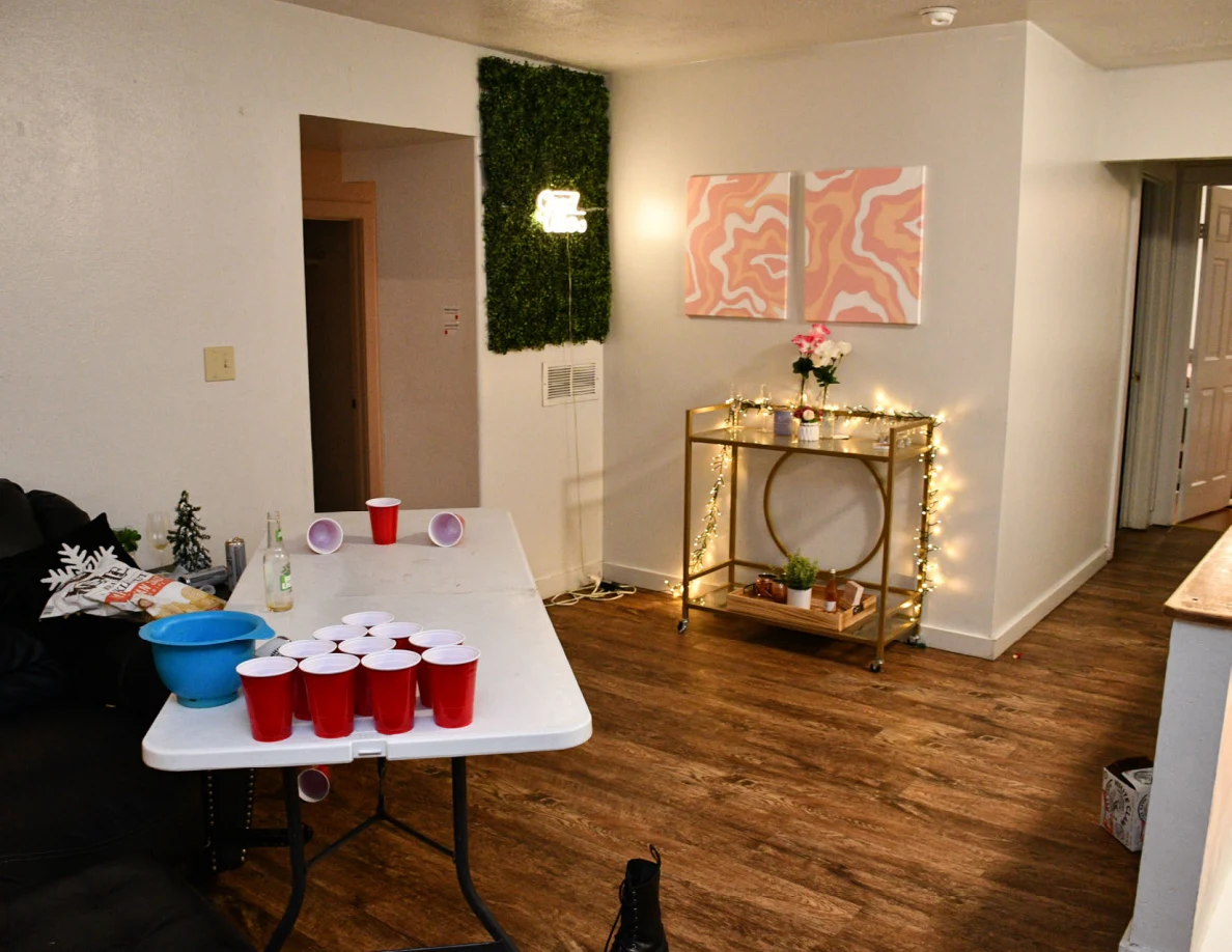 Cup-pong table with red cups and a blue bowl beside a dark couch and gold drinks cart.