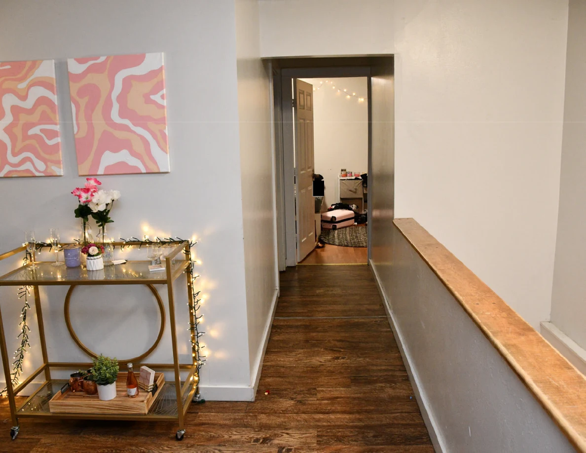 Gold drinks cart below pink wall art beside a hallway and wooden stair rail.