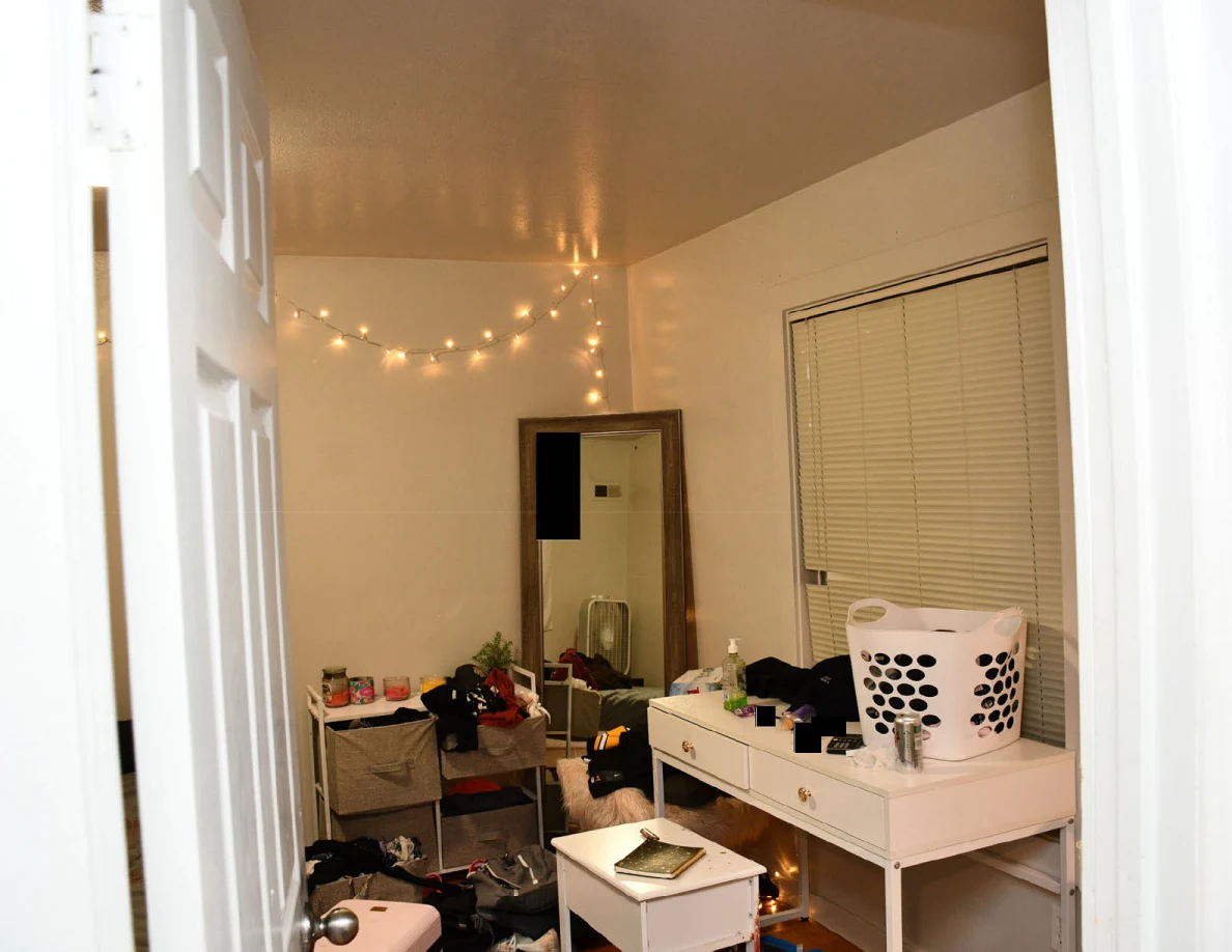 Bedroom desk with laundry basket beside a tall mirror, window blinds and string lights.