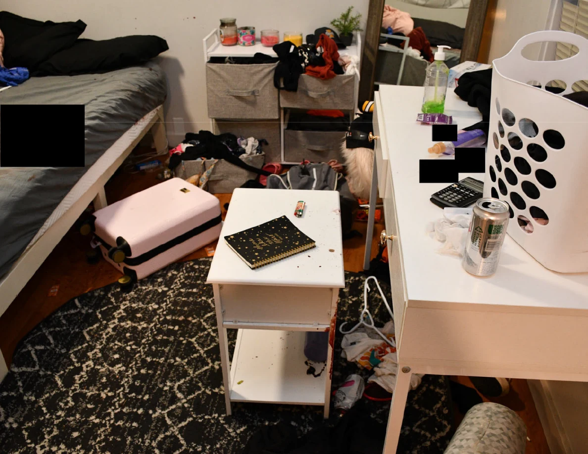 White desk with laundry basket and drink cans beside a small notebook table, patterned rug and pink suitcase.