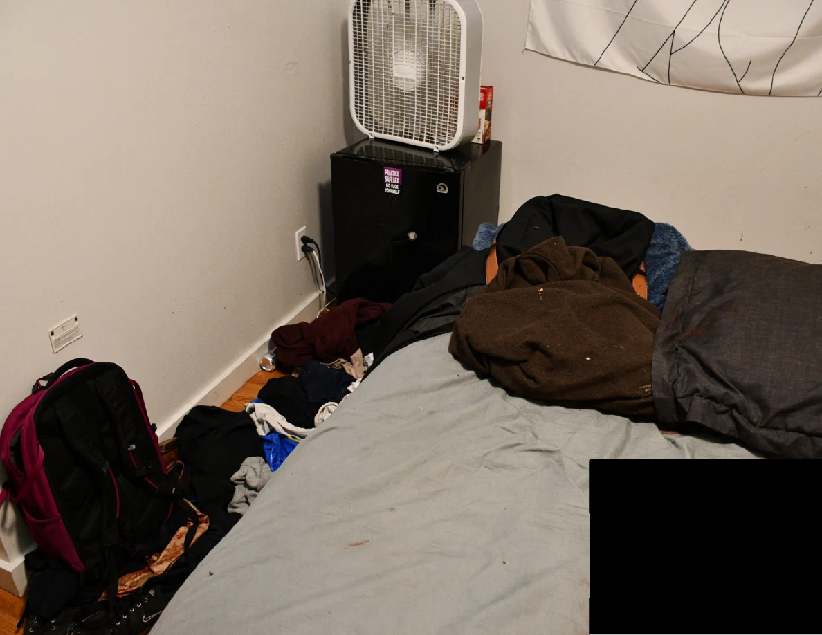 Bed with dark bedding and a brown bag beside a box fan, dark mini fridge and backpack, partly redacted.