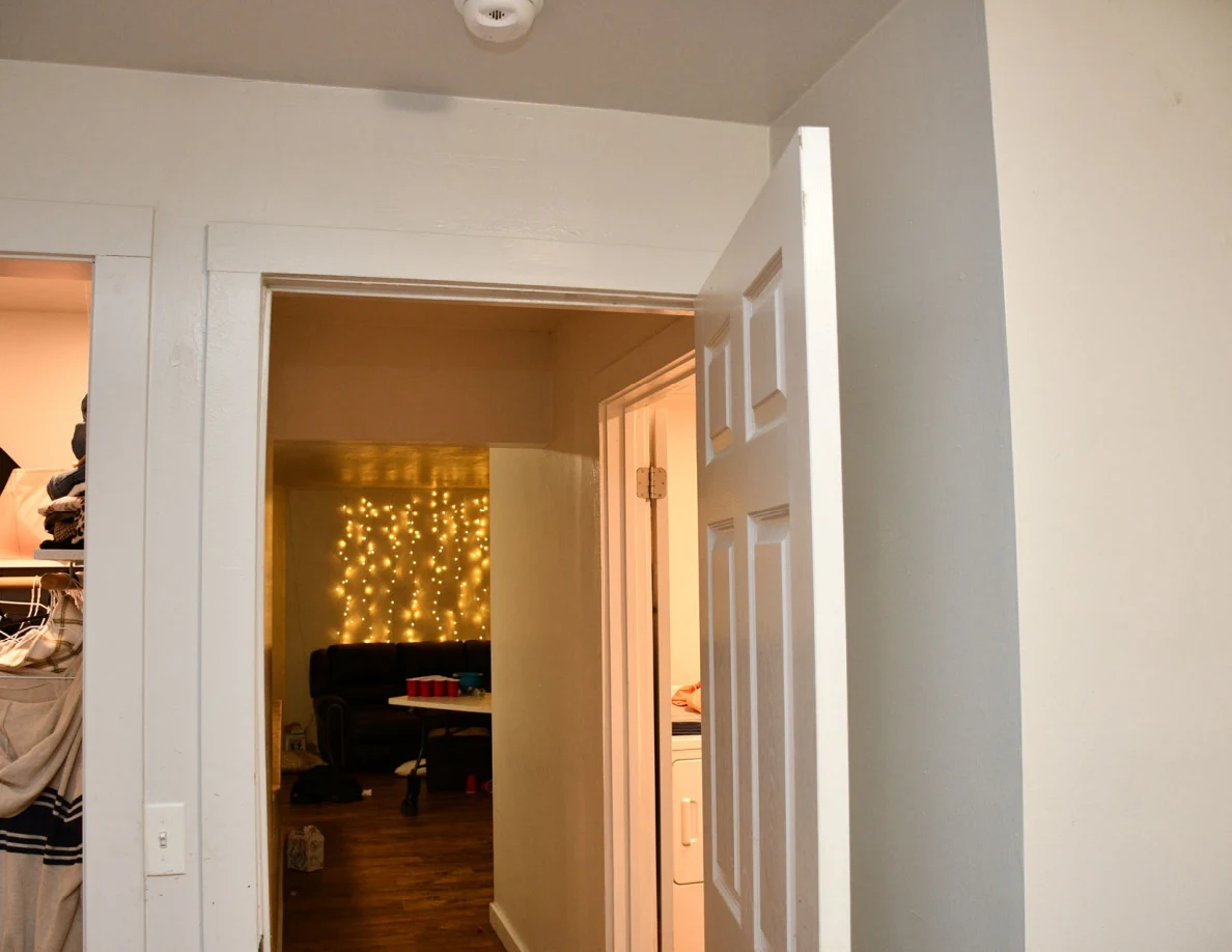 Open bedroom door looking toward a hallway and distant wall of string lights.