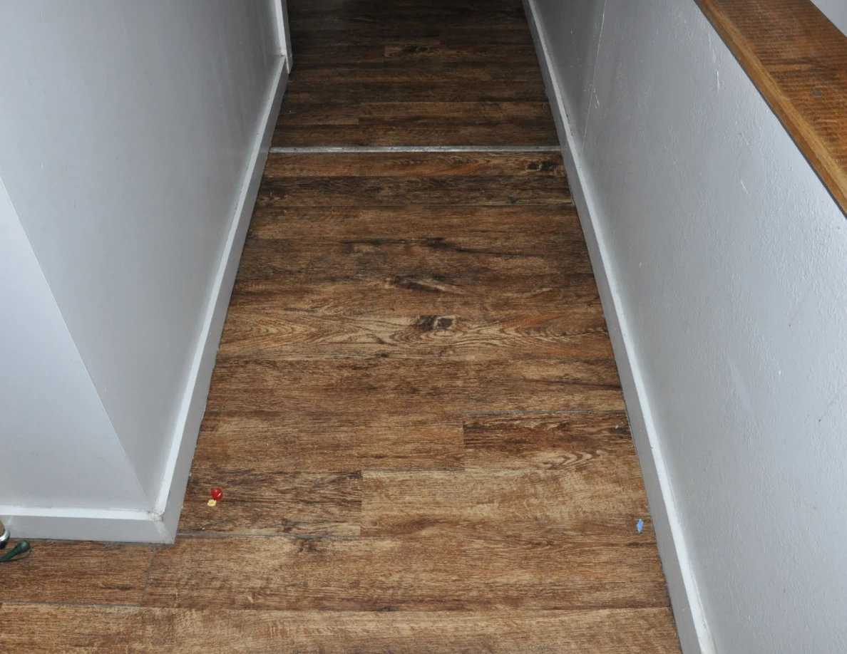 Wood-pattern hallway floor with a narrow threshold strip beside a stair rail.