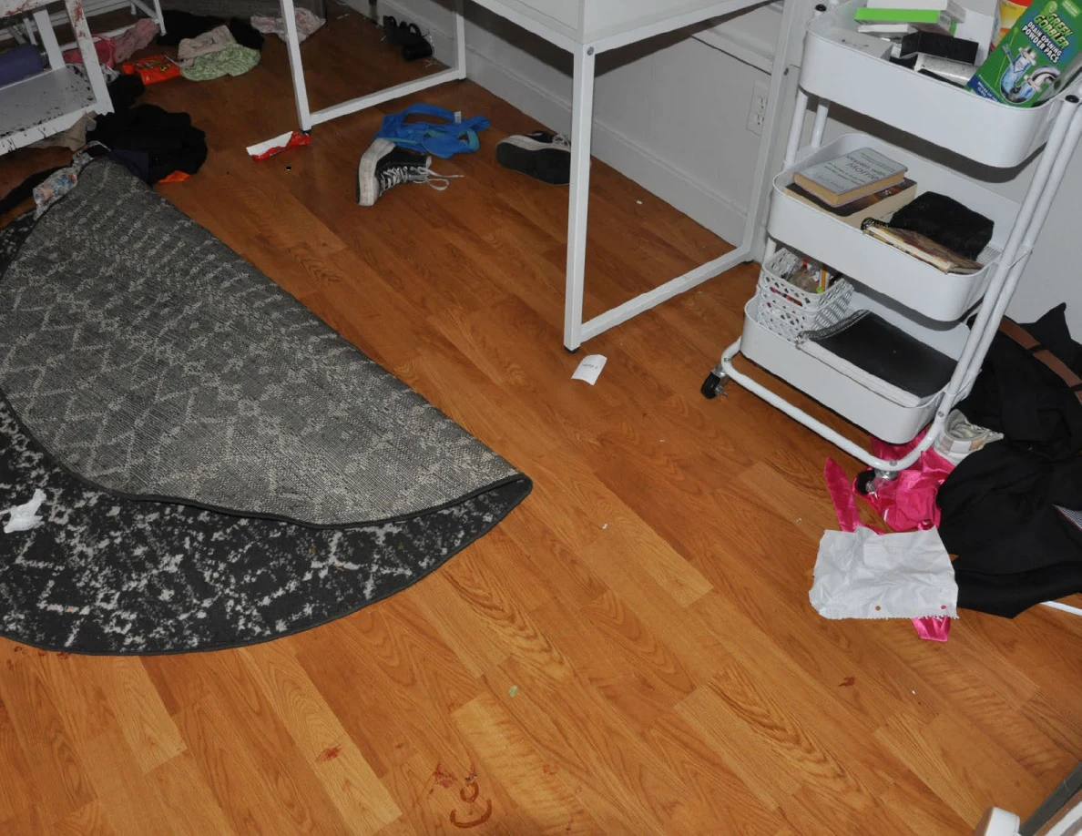 Folded patterned rug beside desk legs, a rolling cart, footwear and clothing.
