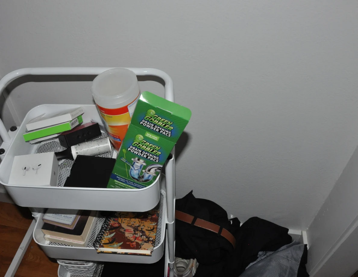 White rolling cart with cleaning products and books beside dark clothing.