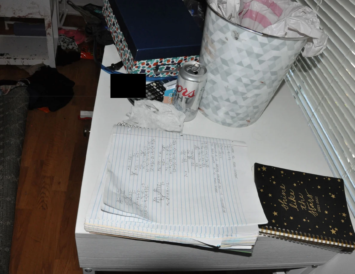 Open handwritten notebook beside a drink can and patterned wastebasket on a white desk.