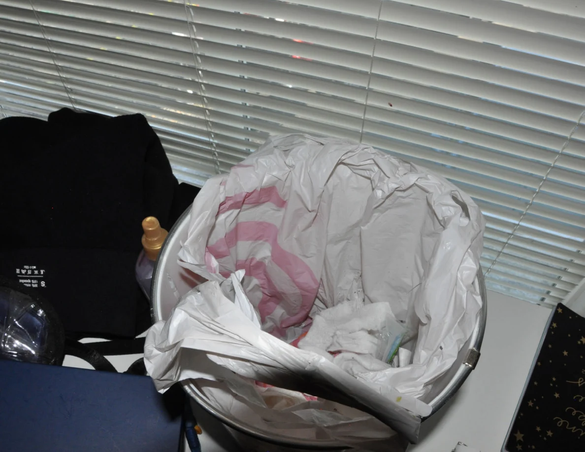 Patterned wastebasket lined with shopping bags beneath closed blinds.