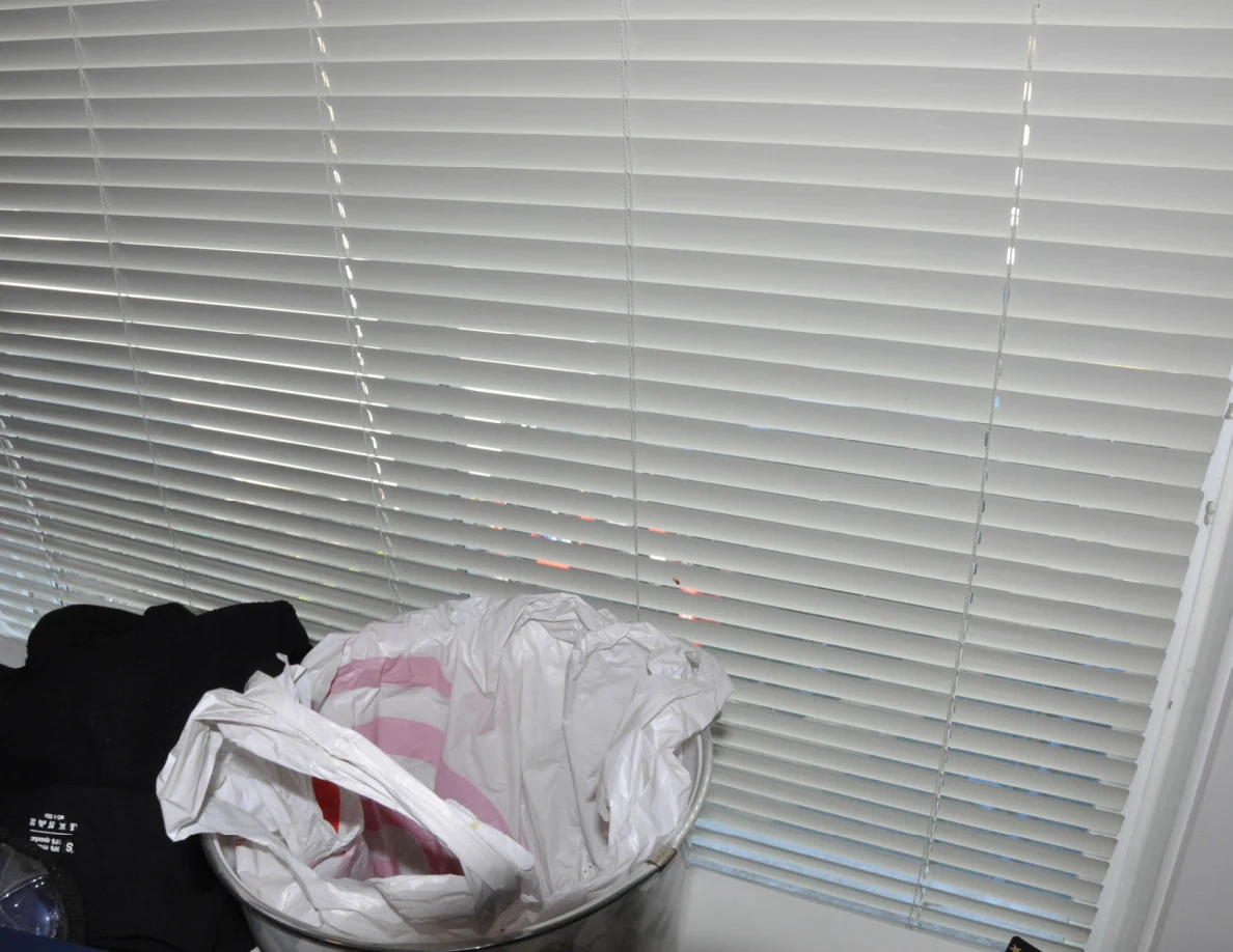 Closed blinds above a wastebasket containing shopping bags.