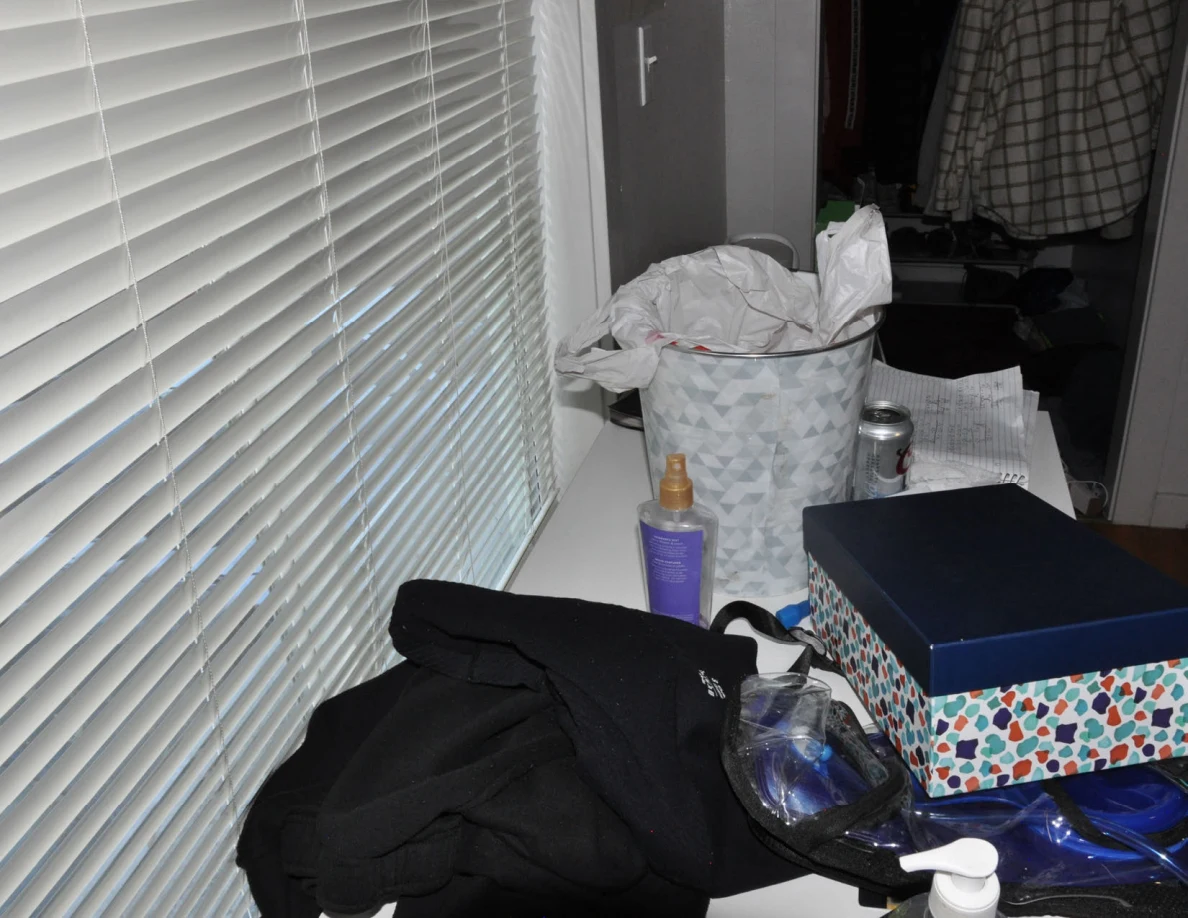Desk beneath blinds with clothing, bottles, patterned box and lined wastebasket.