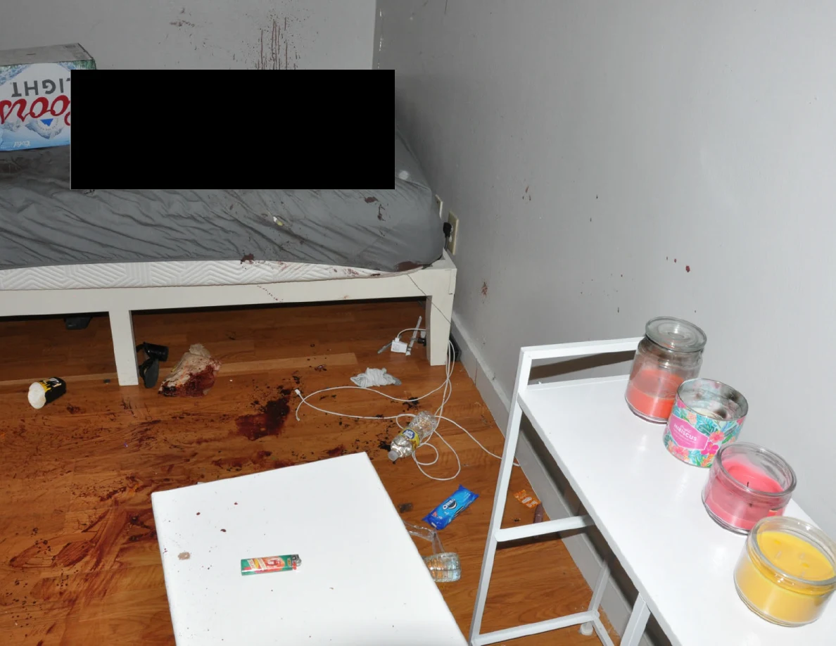 Partly redacted bed beside reddish floor and wall marks, cables and white furniture.