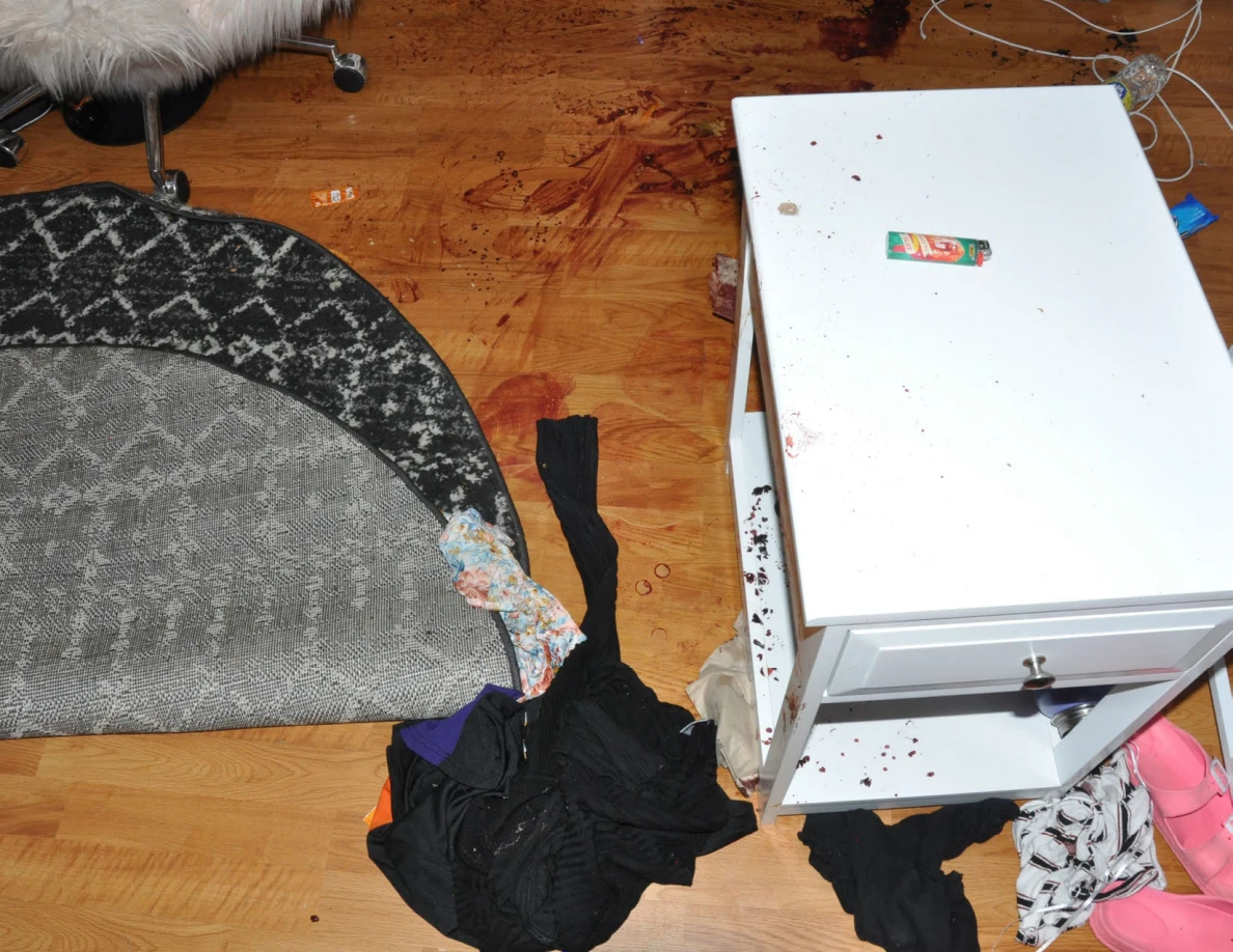 White bedside table beside a folded rug, clothing and reddish floor marks.