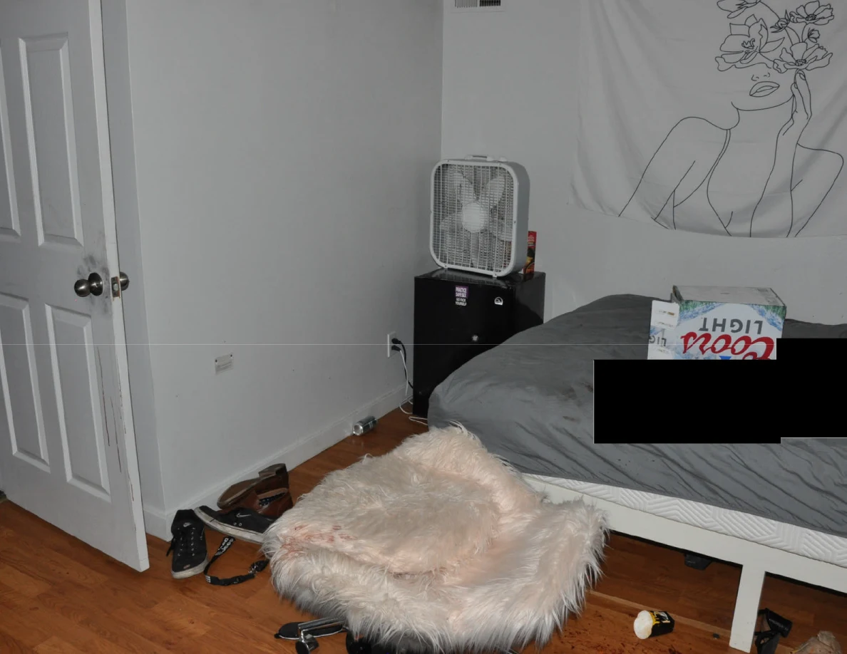 Fluffy chair beside a partly redacted bed and box fan on a mini fridge.
