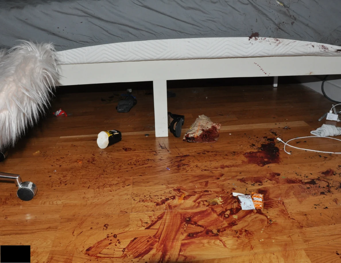 White bed frame and gray sheet above reddish floor marks, scattered items and cables.
