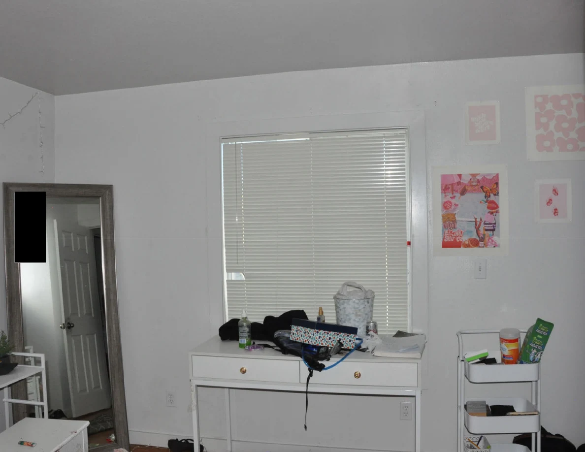 White desk beneath closed blinds beside pink wall prints and a rolling cart.