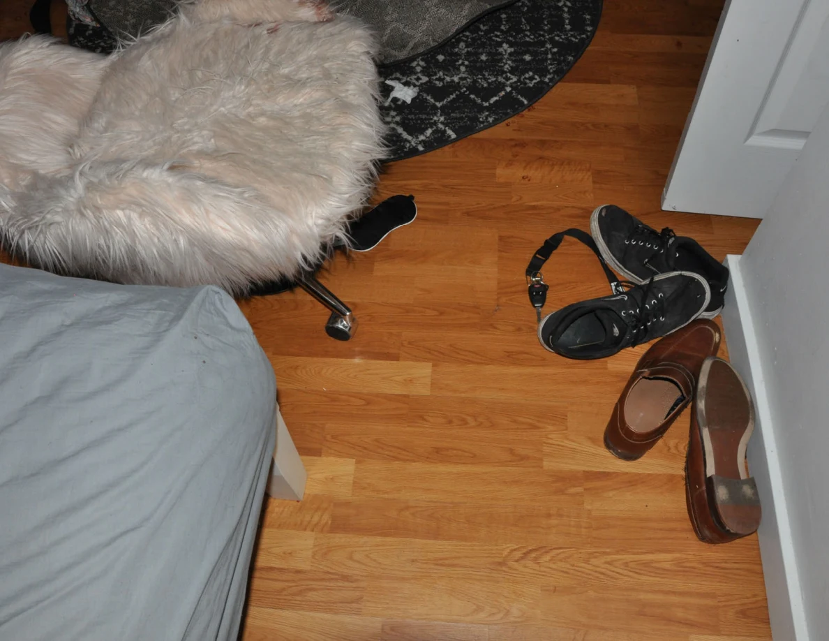 Footwear and a strap on the floor beside a fluffy chair and bed.