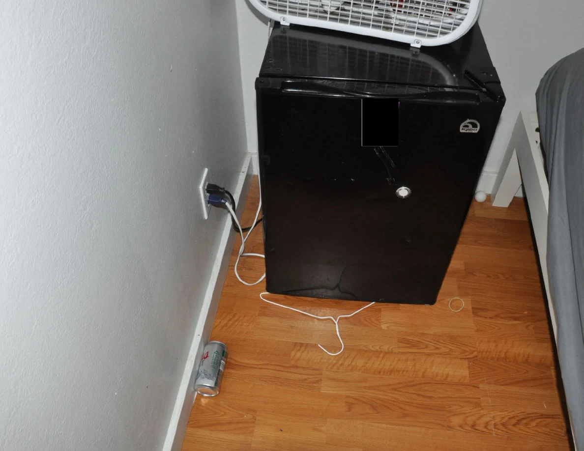 Dark mini fridge with a box fan on top and white cable on the floor.