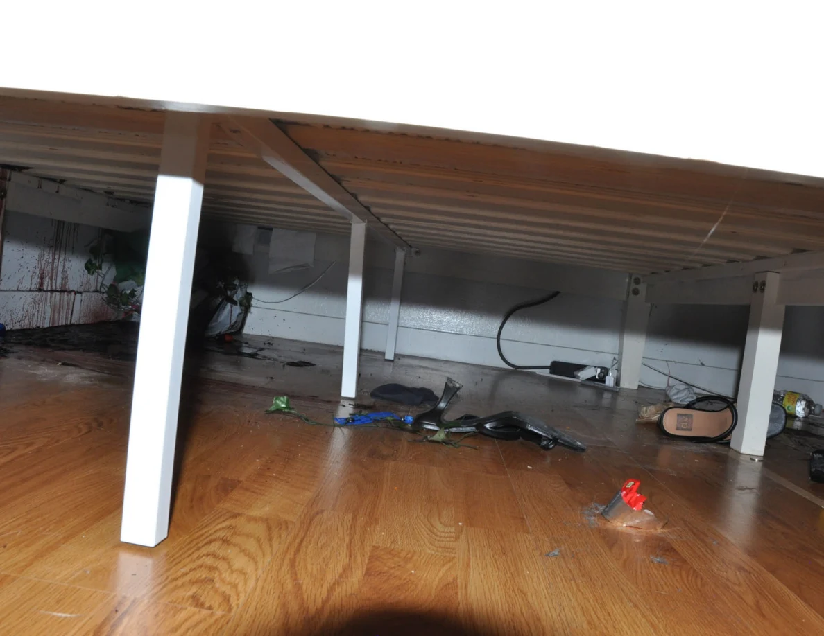 Low view beneath a white bed frame showing slats, footwear, cables and scattered items.