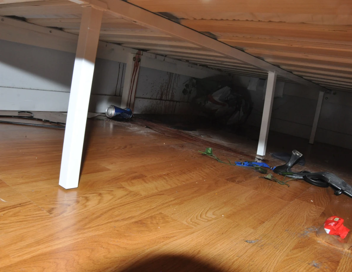 Low view under a bed showing white supports, reddish wall streaks and scattered objects.
