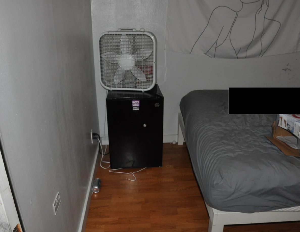 Box fan on a dark mini fridge beside a partly redacted bed.
