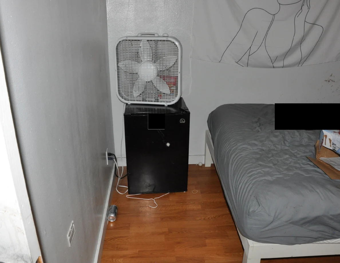 Mini fridge and box fan beside a gray-covered bed and white cable.