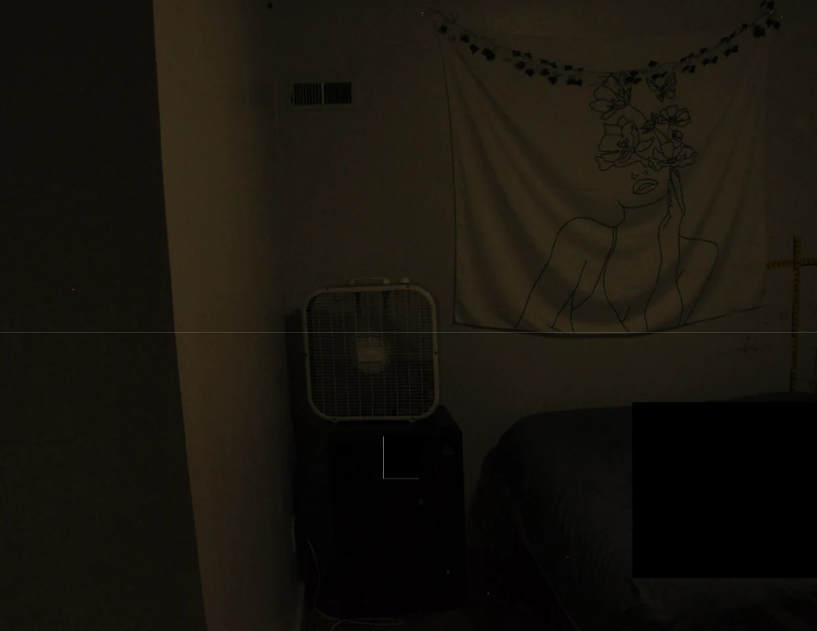 Dim view of a box fan below a floral wall hanging beside redacted bedding.