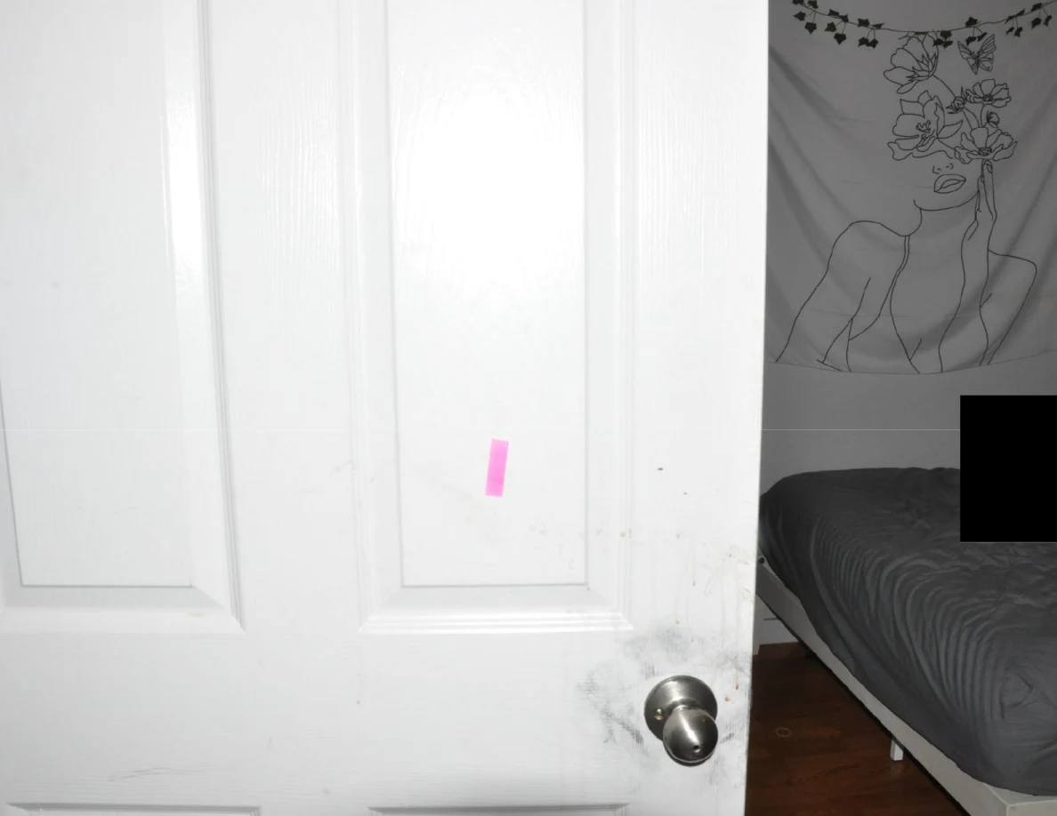Open door with pink tape beside a floral wall hanging and partly redacted bed.