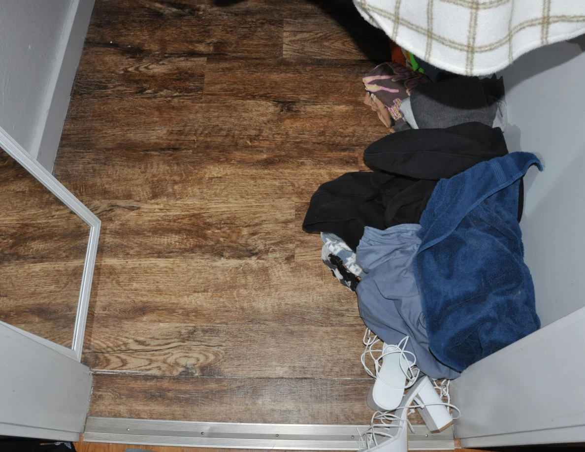Closet floor with piled clothing and white strappy footwear.