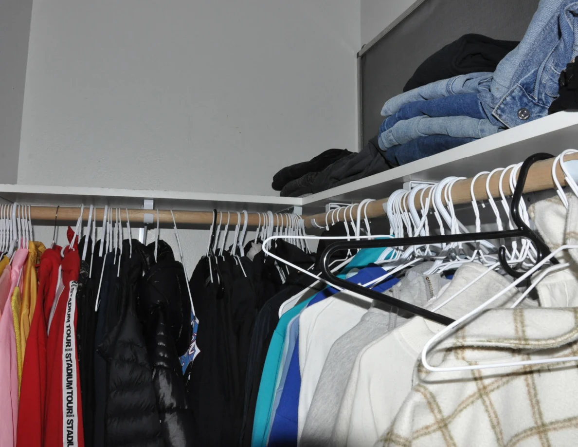 Closet rails with hangers and garments beneath folded jeans on a shelf.