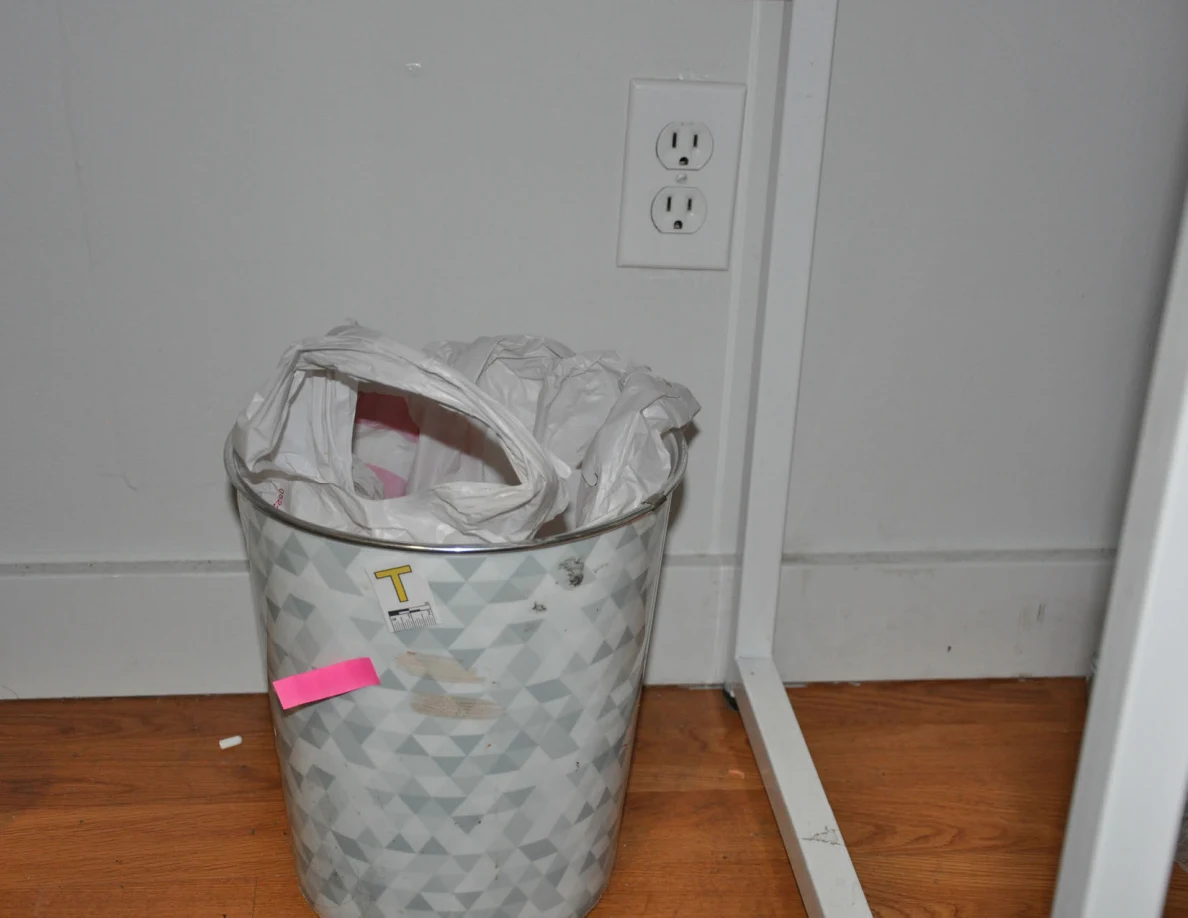 Patterned wastebasket with scale label T and pink tape below a wall outlet.