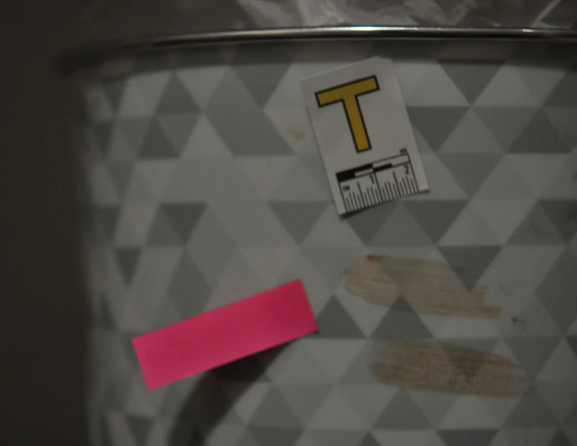 Close view of pale smears on a patterned wastebasket beside label T and pink tape.