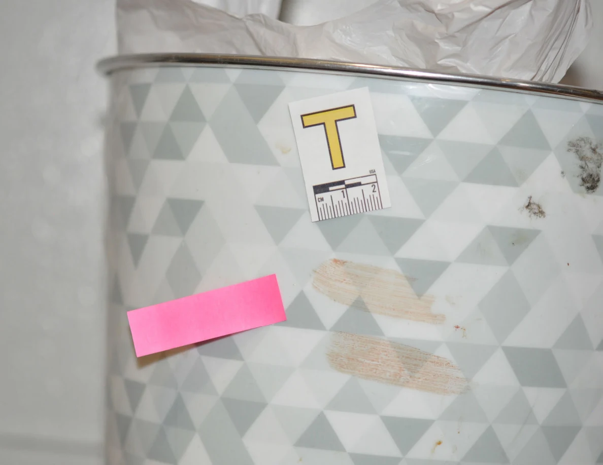 Close view of pale smears on a patterned wastebasket beside label T and pink tape.