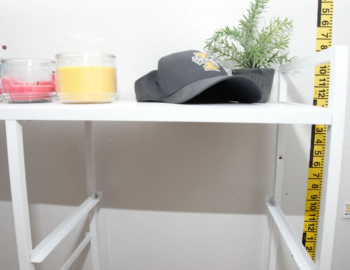 Candles, a dark cap and small plant on a white cabinet beside measuring tape.
