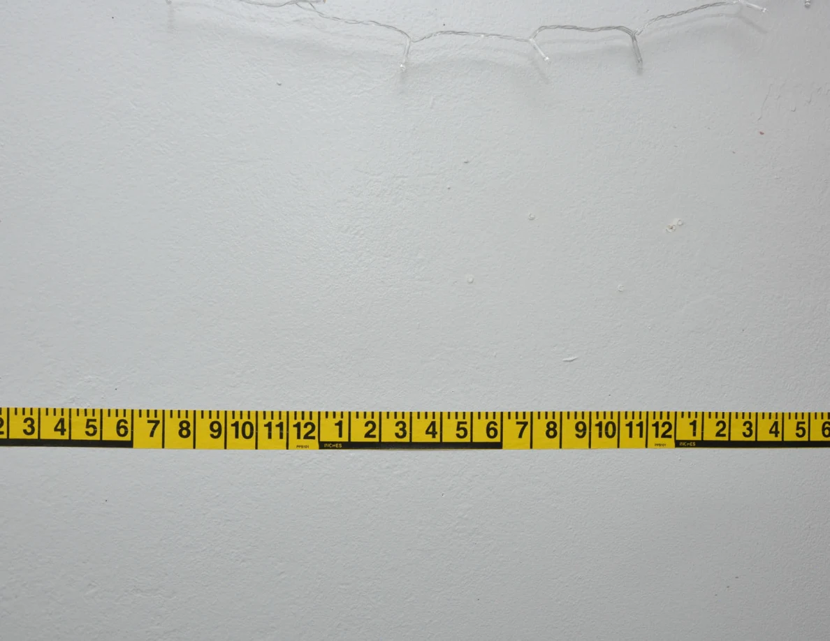 Horizontal yellow measuring tape below string lights on a pale wall.