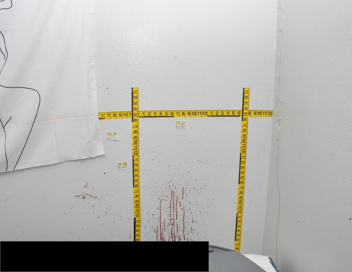 Wall corner with reddish streaks, fabric hanging, crossed yellow measuring tapes and a black redaction below.