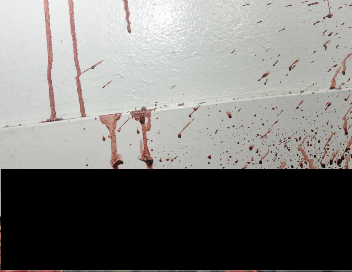 Reddish spots and streaks on a wall and baseboard above a black redaction.