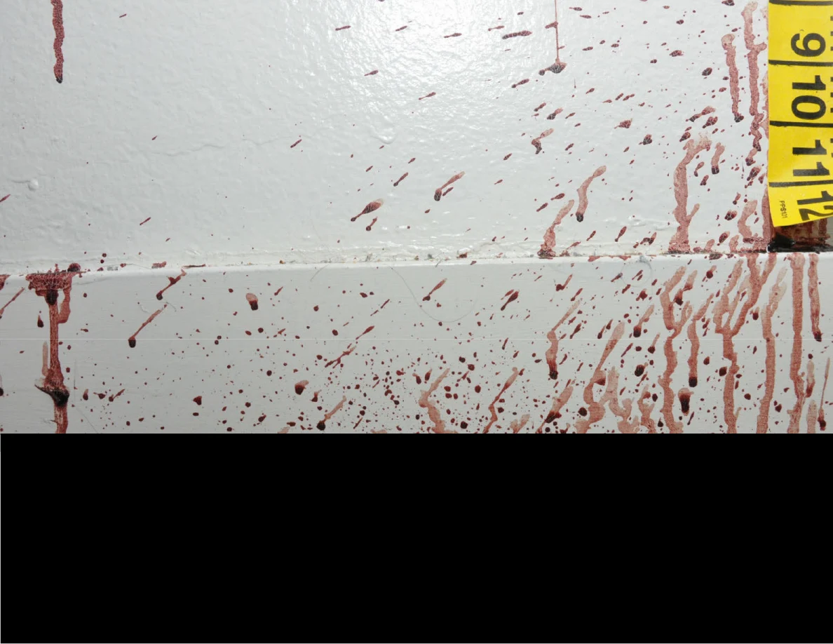 Reddish spots on a wall and baseboard beside yellow measuring tape, above a black redaction.