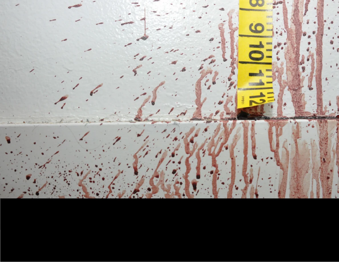 Reddish wall and baseboard streaks beside yellow measuring tape above a black redaction.