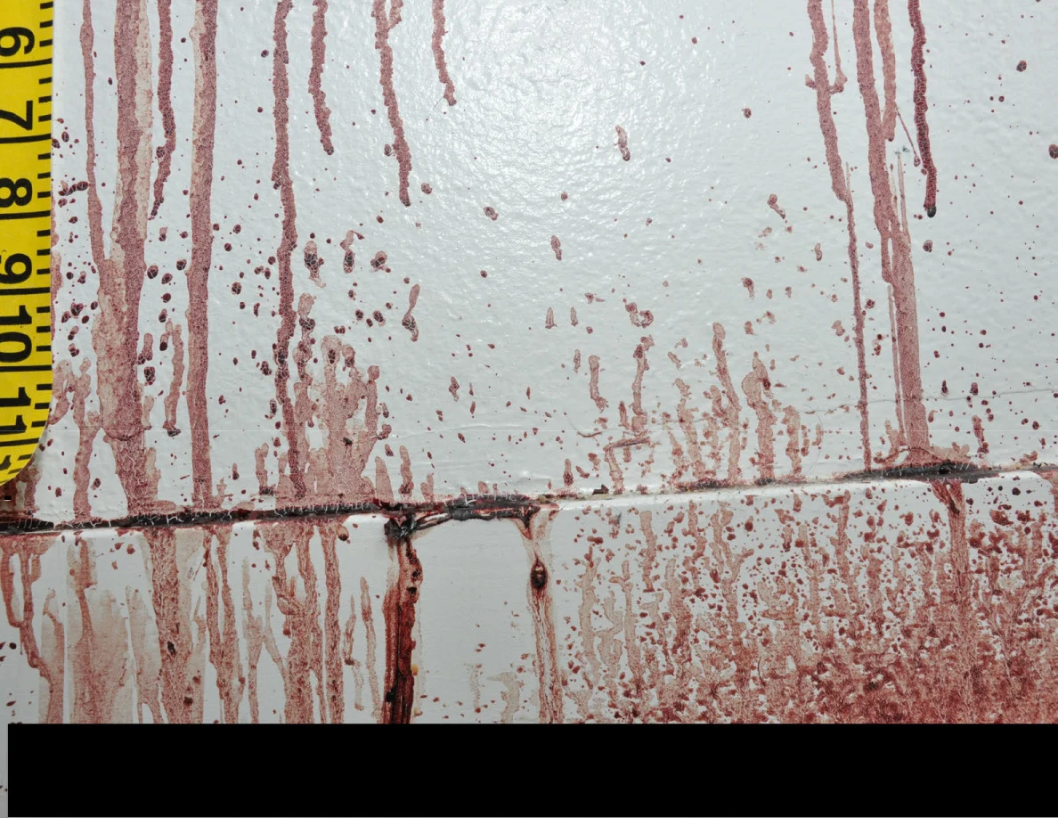 Reddish wall and baseboard streaks beside measuring tape above a black redaction.