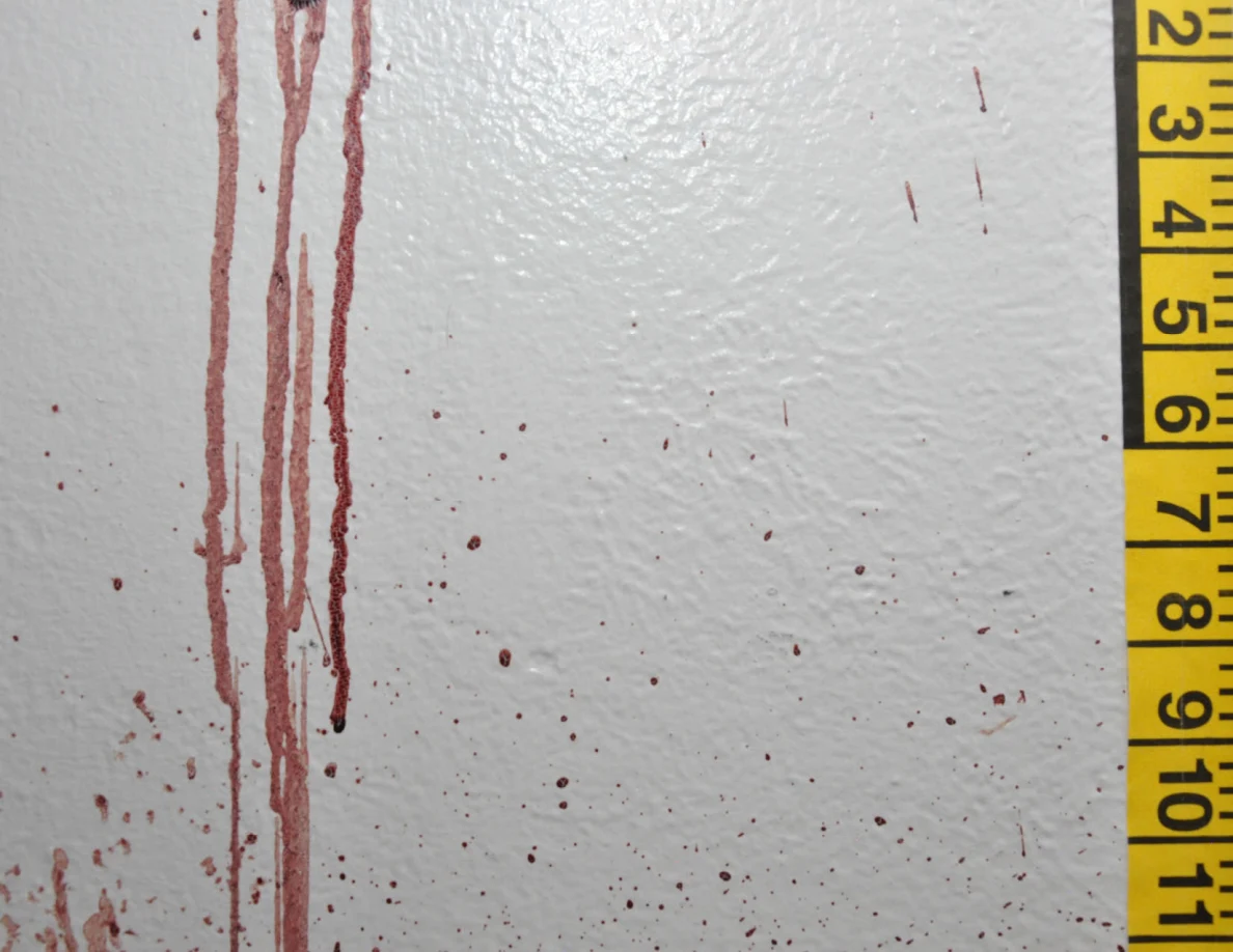 Long reddish streaks and small spots on a white wall beside yellow measuring tape.