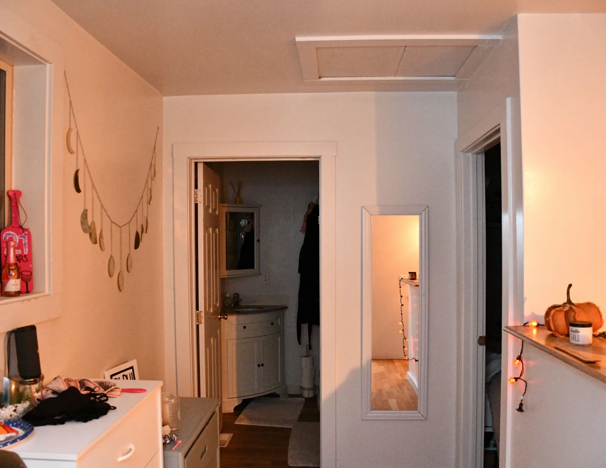 Hallway with wall mirror, white drawers, ceiling hatch and pumpkin decoration beside string lights.