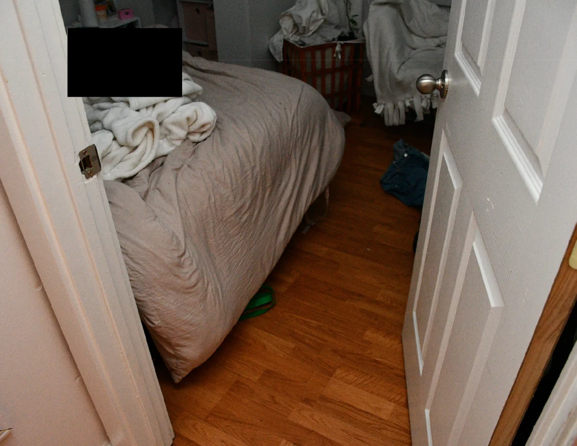 Bed with rumpled bedding beside wood floor and an open white door, with a black redaction.