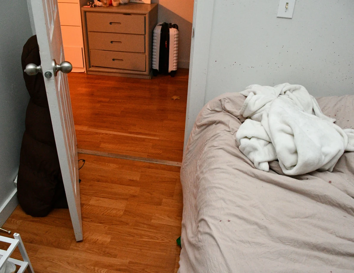 Rumpled white blanket on bed beside open doorway, wood floor and hall drawers.