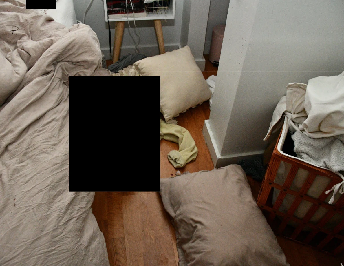 Pillows, bedding and a laundry basket beside wood flooring and black redactions.
