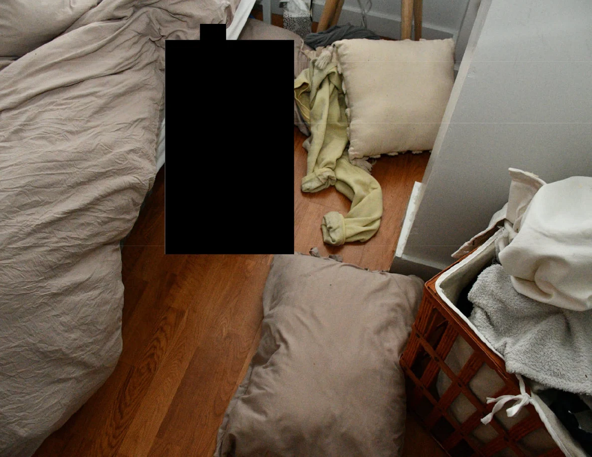 Pillows and pale fabric on wood floor beside bed and laundry basket, with a black redaction.