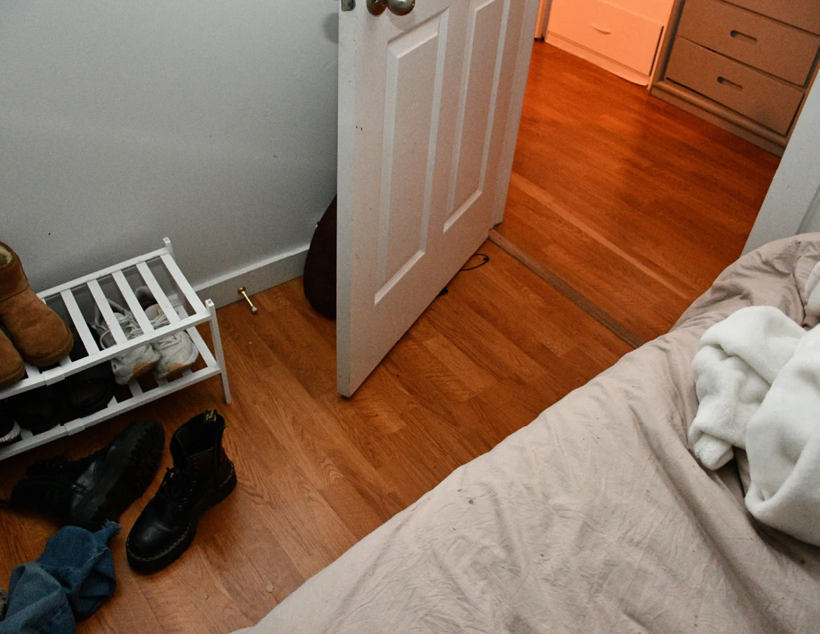 White shoe rack and black boots on wood floor beside bed and an open bedroom door.