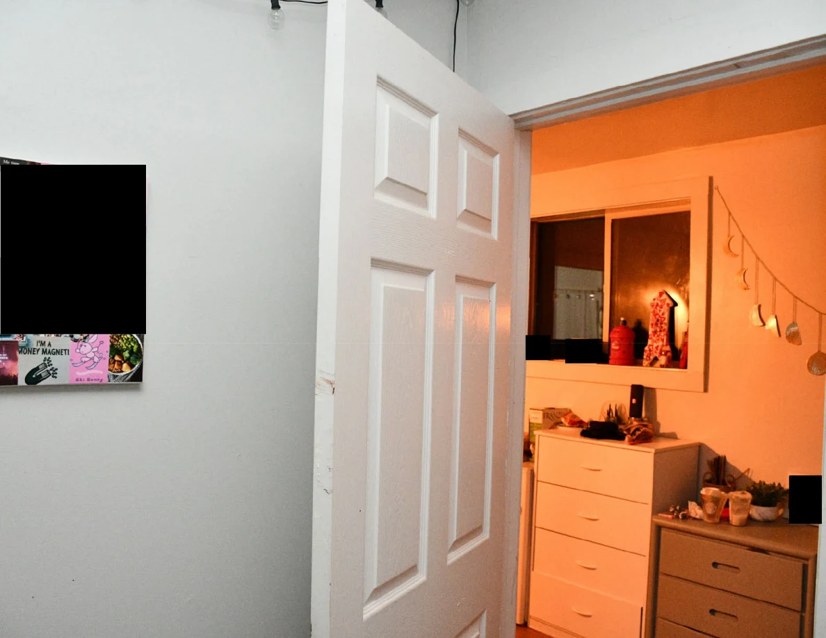 Open white bedroom door looking toward hall drawers and hanging decorations, with a black redaction on wall art.