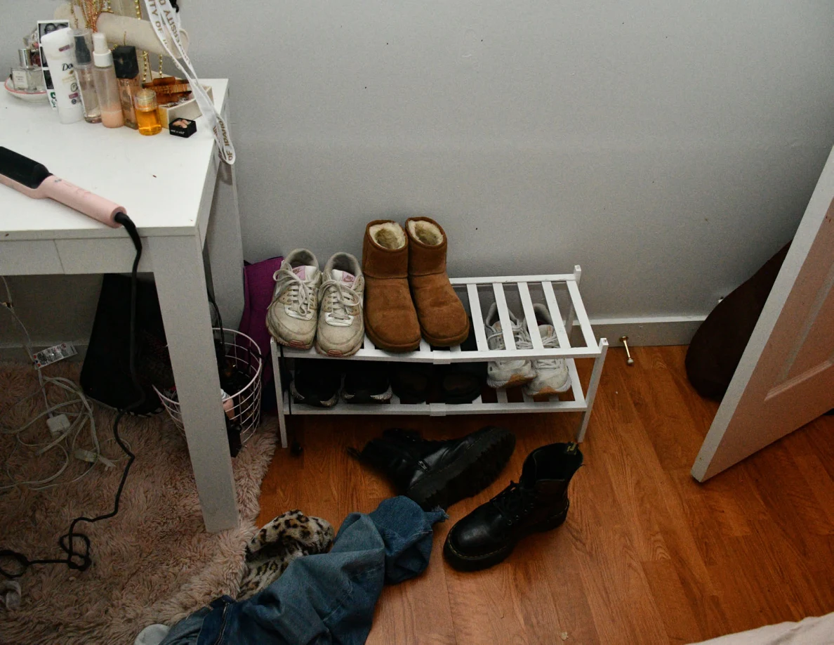 Shoe rack with tan boots and sneakers beside white desk, cosmetics and clothing on wood floor.