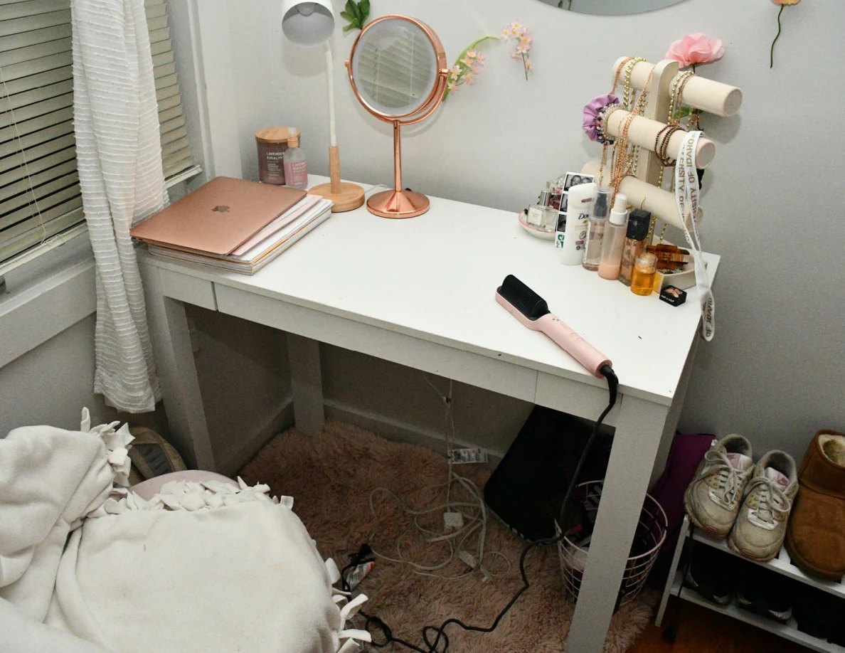 White desk with closed rose-colored laptop, tabletop mirror, jewelry stand, cosmetics and hair-styling tool.