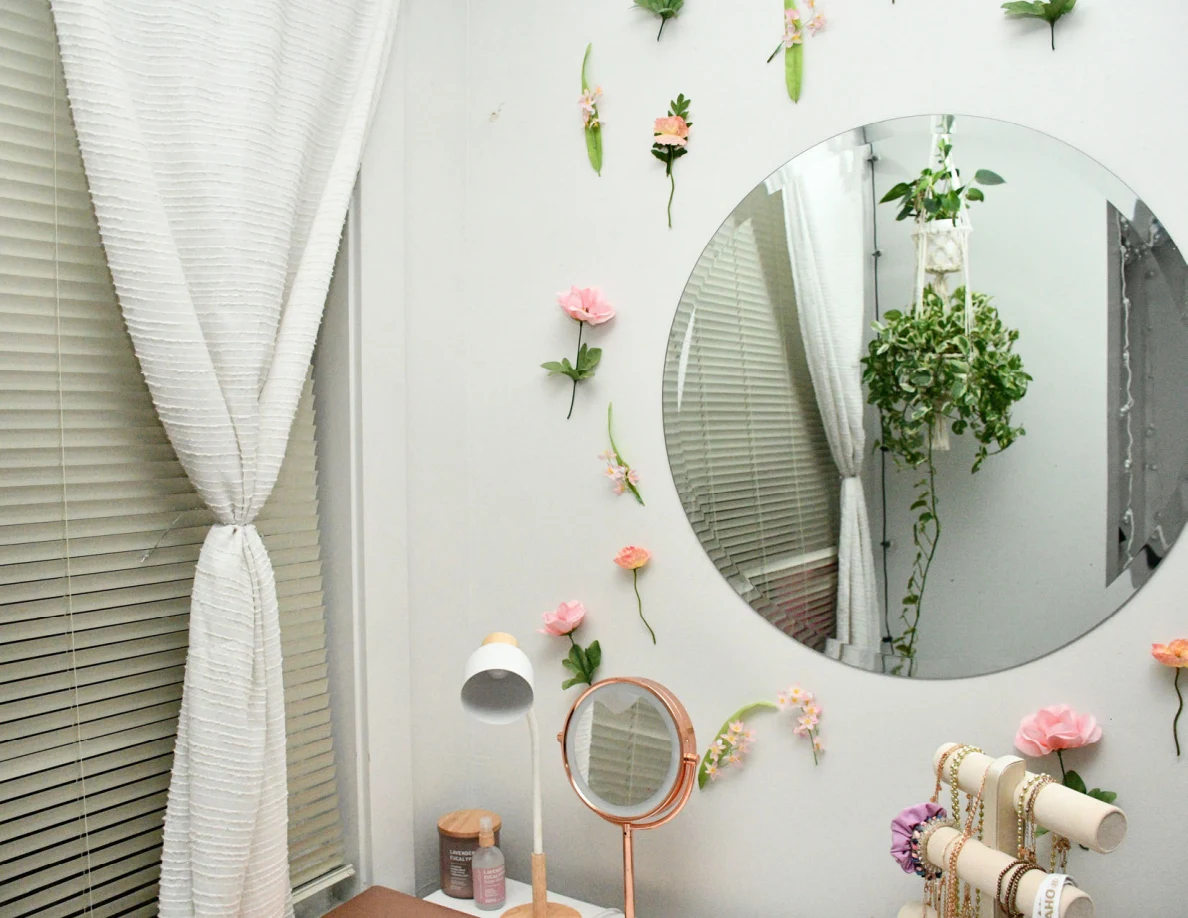 Round wall mirror surrounded by flowers beside curtains and tabletop mirror.
